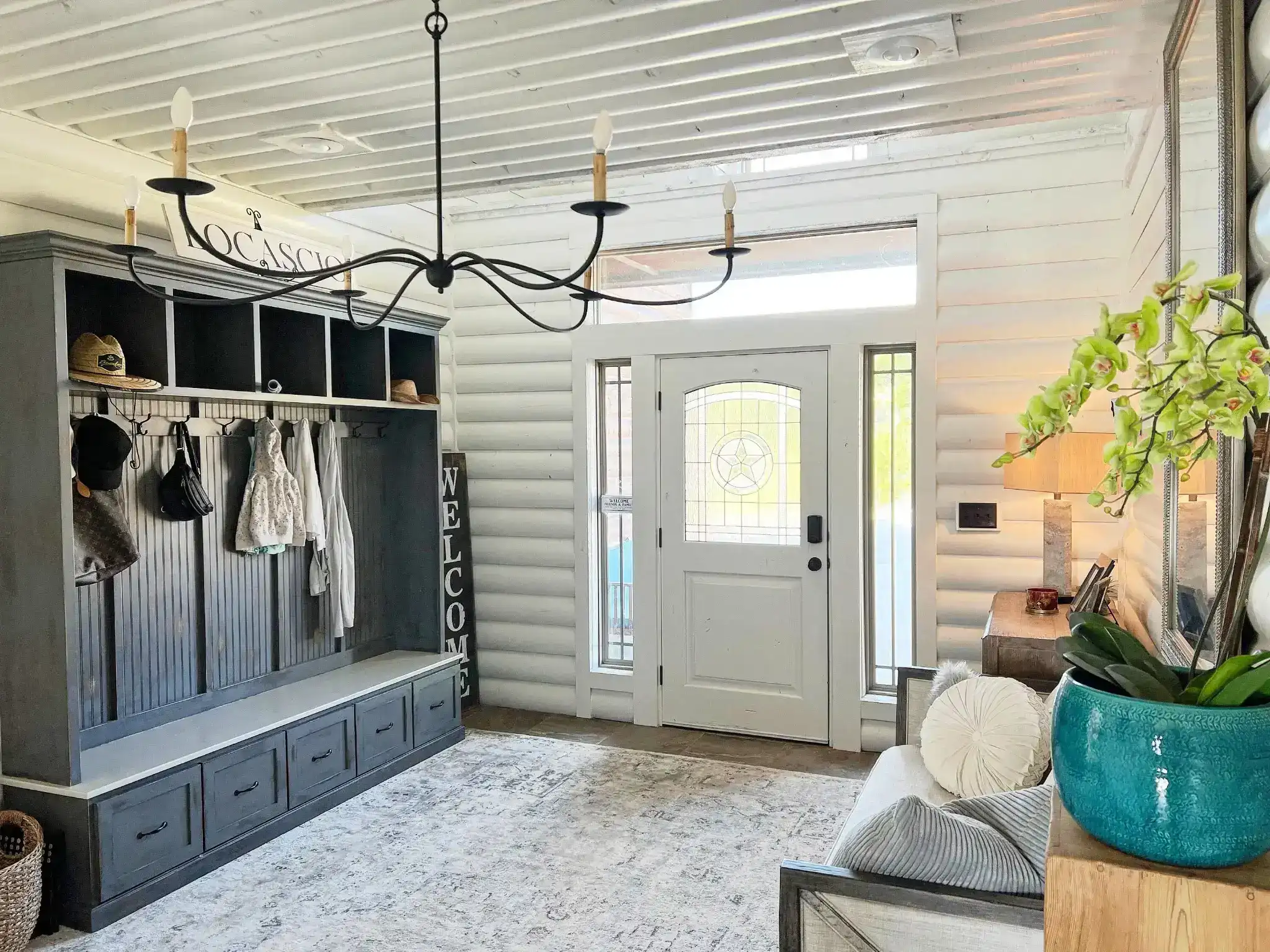Bright farmhouse-style mudroom with a built-in dark gray storage bench featuring drawers, open cubbies, and wall hooks for coats and hats.