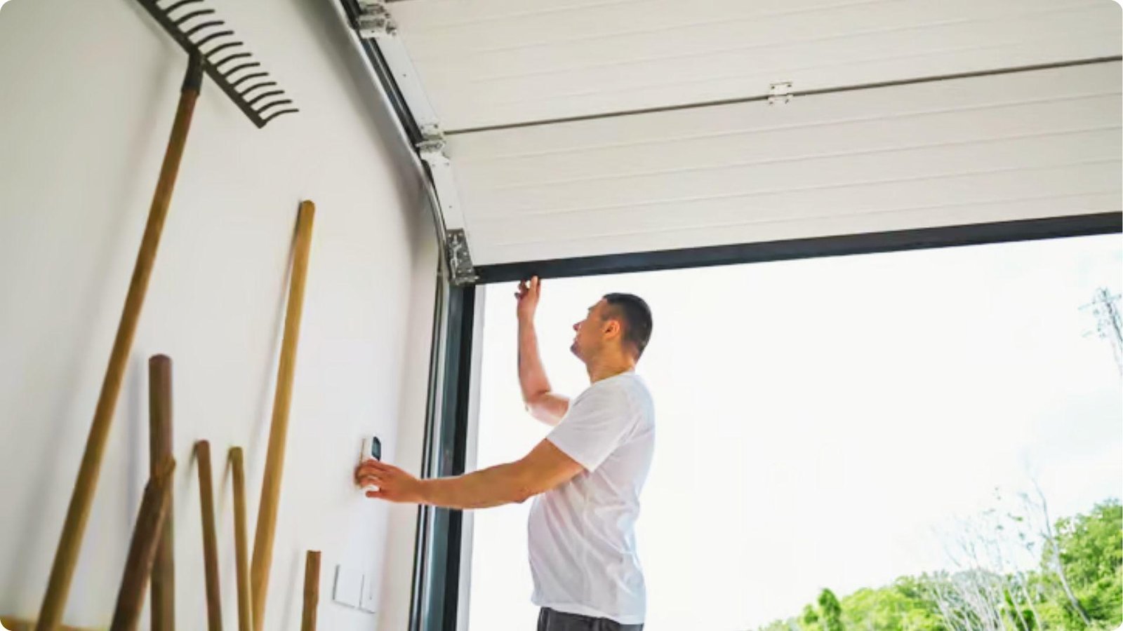 A man in a white t-shirt inspects a partially open garage door while pressing the wall opener.