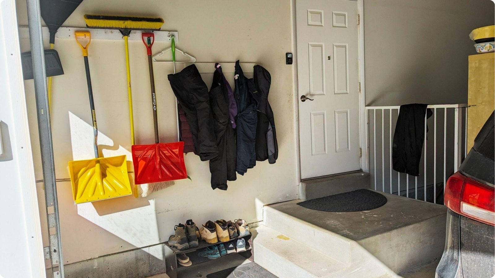 Practical garage mudroom area with wall-mounted shovels, brooms, and coats near the home’s interior door.