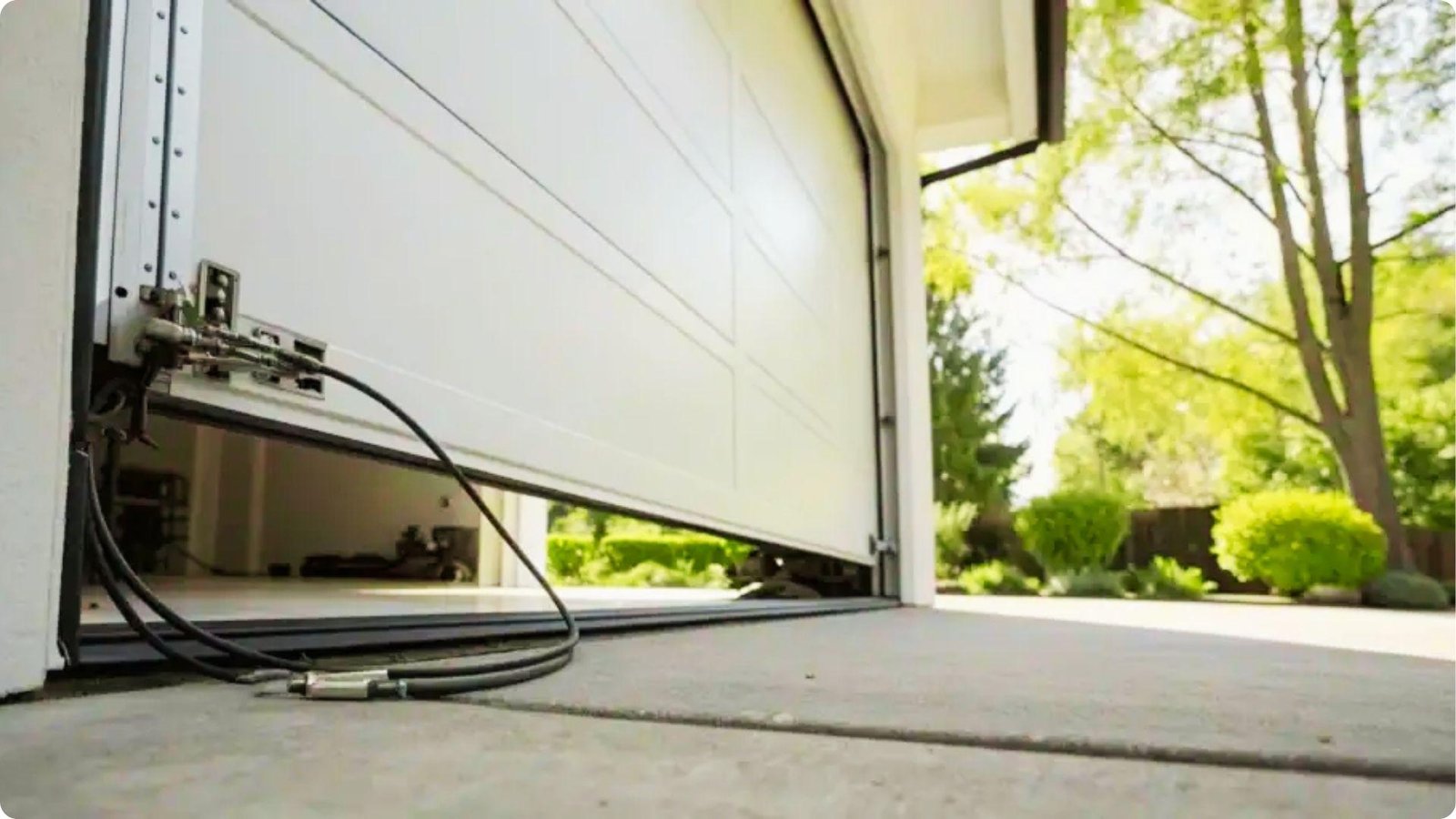 A garage door lowered unevenly with a black lift cable detached and lying on the driveway.