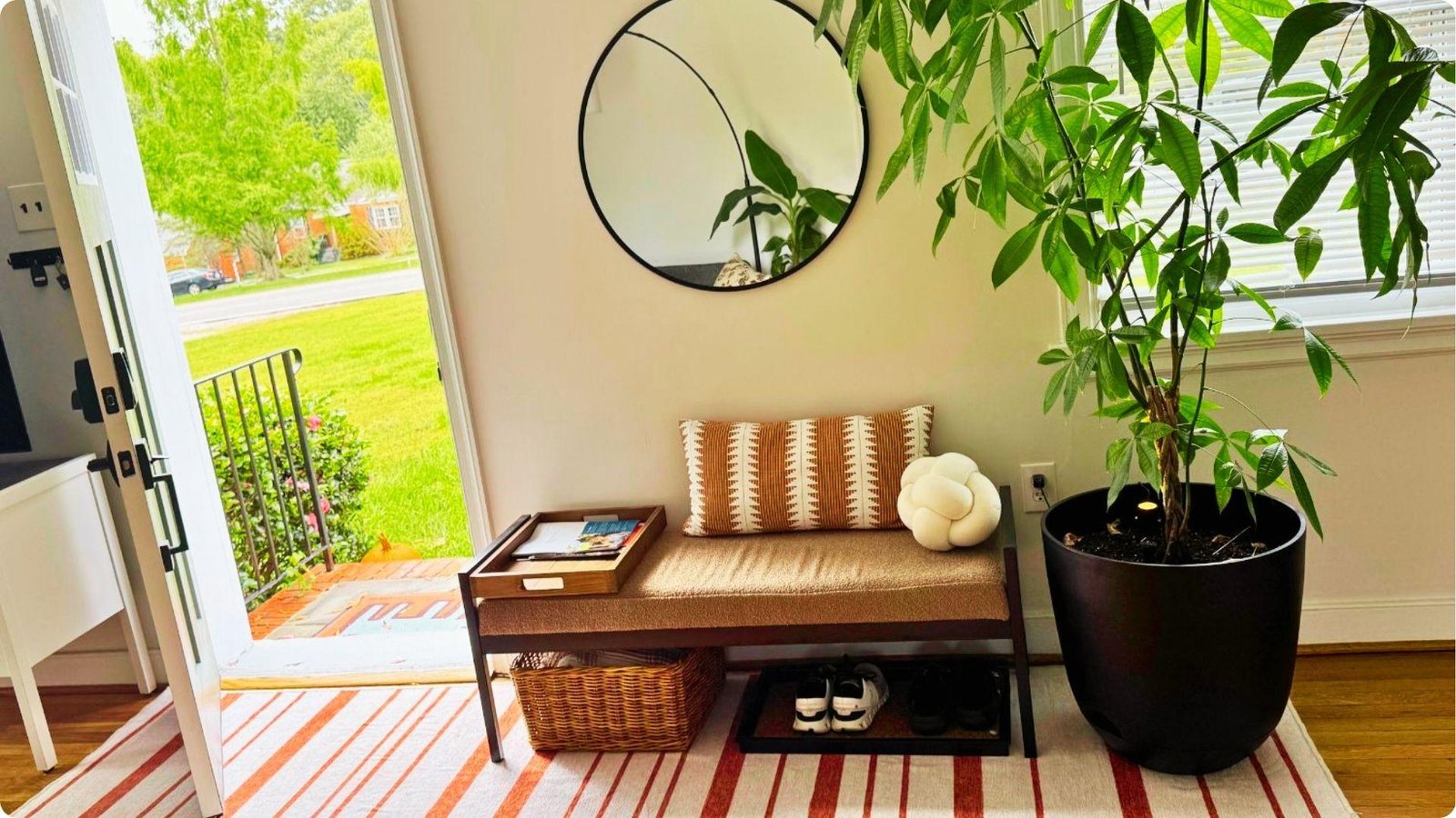 An entry bench with striped pillow sits by an open front door, topped with a round mirror and flanked by a tall plant.