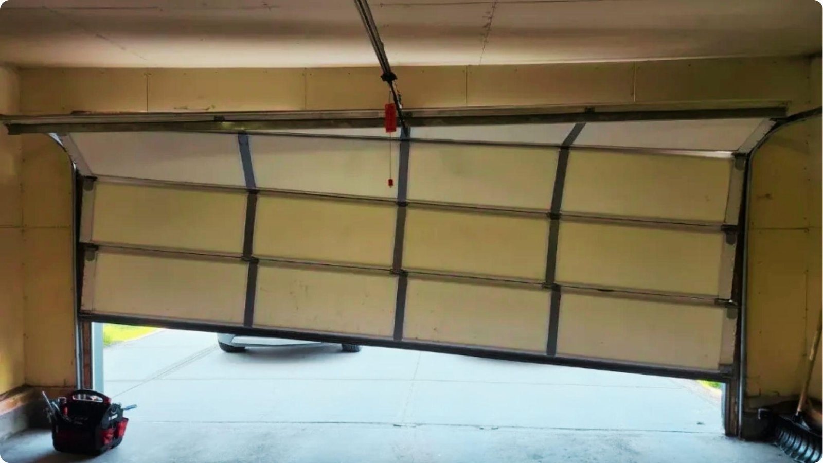 A garage door hangs crooked and uneven in its tracks with a tool bag on the floor.