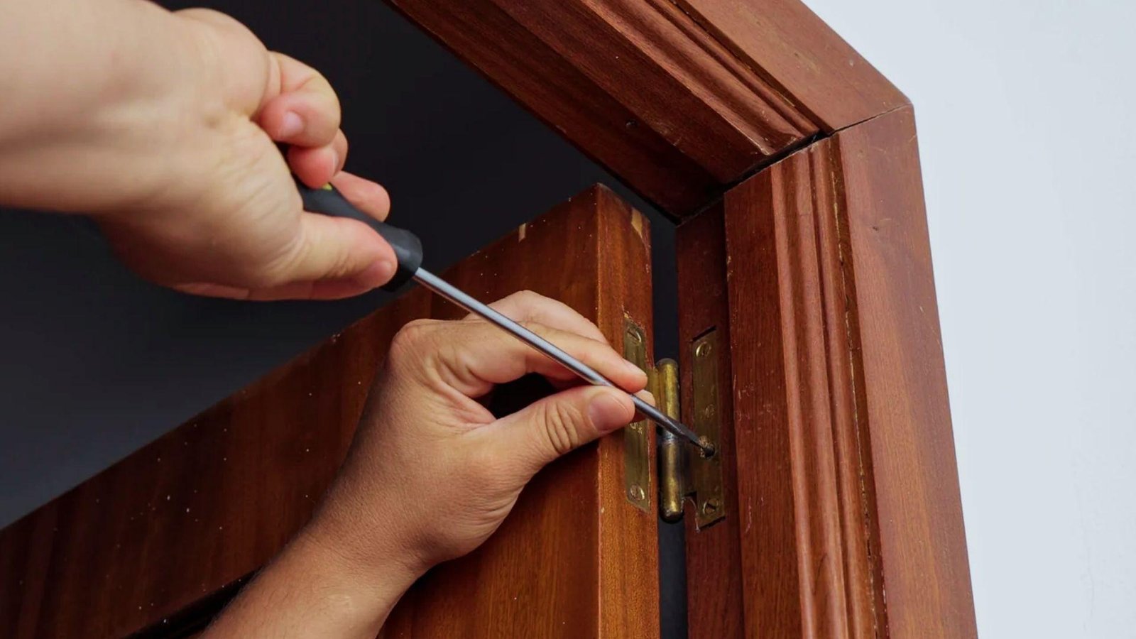Hands using screwdriver to tighten loose hinge screws on wooden door frame to fix sticking door without removal